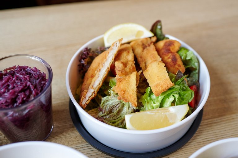 Ein bunter Salat mit einem geschnittenen Schnitzel steht in einer To-Go-Schüssel auf einem Holztisch. Links neben dem Salat steht ein Glas mit Rotkohl.