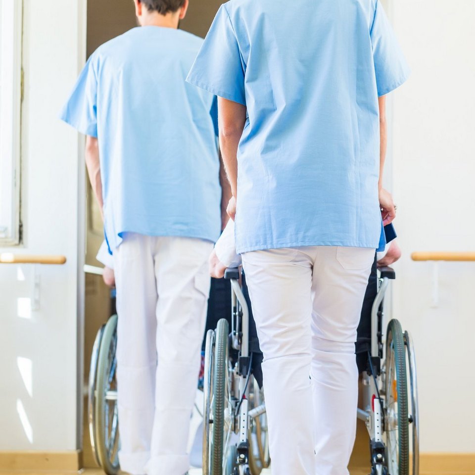 Zwei Krankenschwestern schieben Senioren im Rollstuhl durch ein Pflegeheim.
