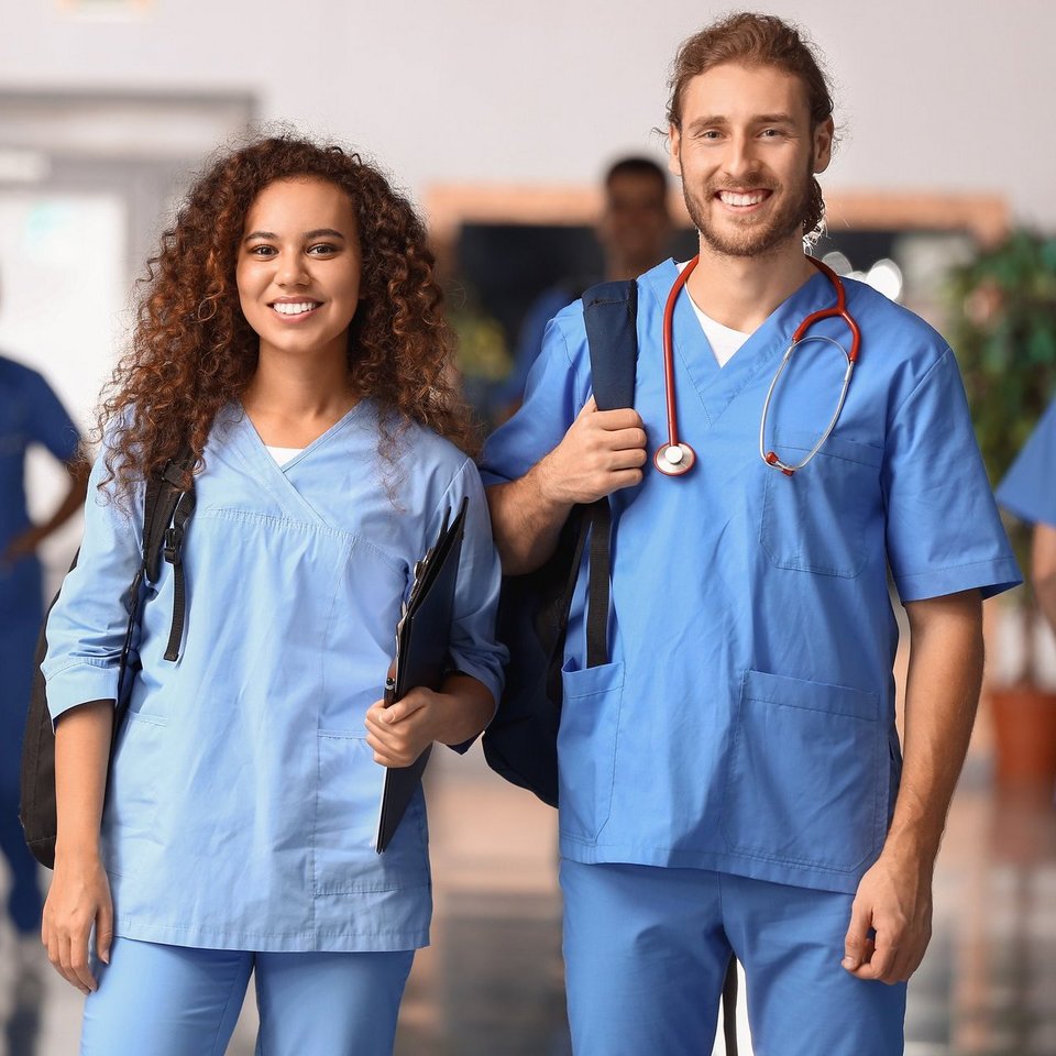 Zwei Pfleger-Auszubildende stehen in einem Gang in dem im Hintergrund mehrere Auszubildende zu erkennen sind.